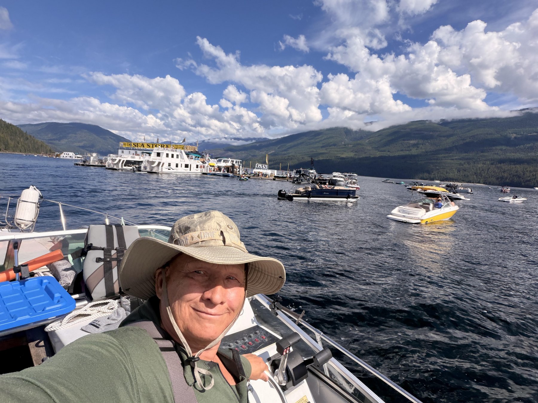 On a boat on Shuswap Lake with the Sea Store houseboat and other boats on the water behind, forested mountains and blue sky beyond