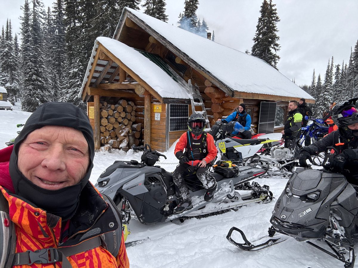 Group of snowmobilers with their sleds gathered outside the snow-covered Owlhead log cabin