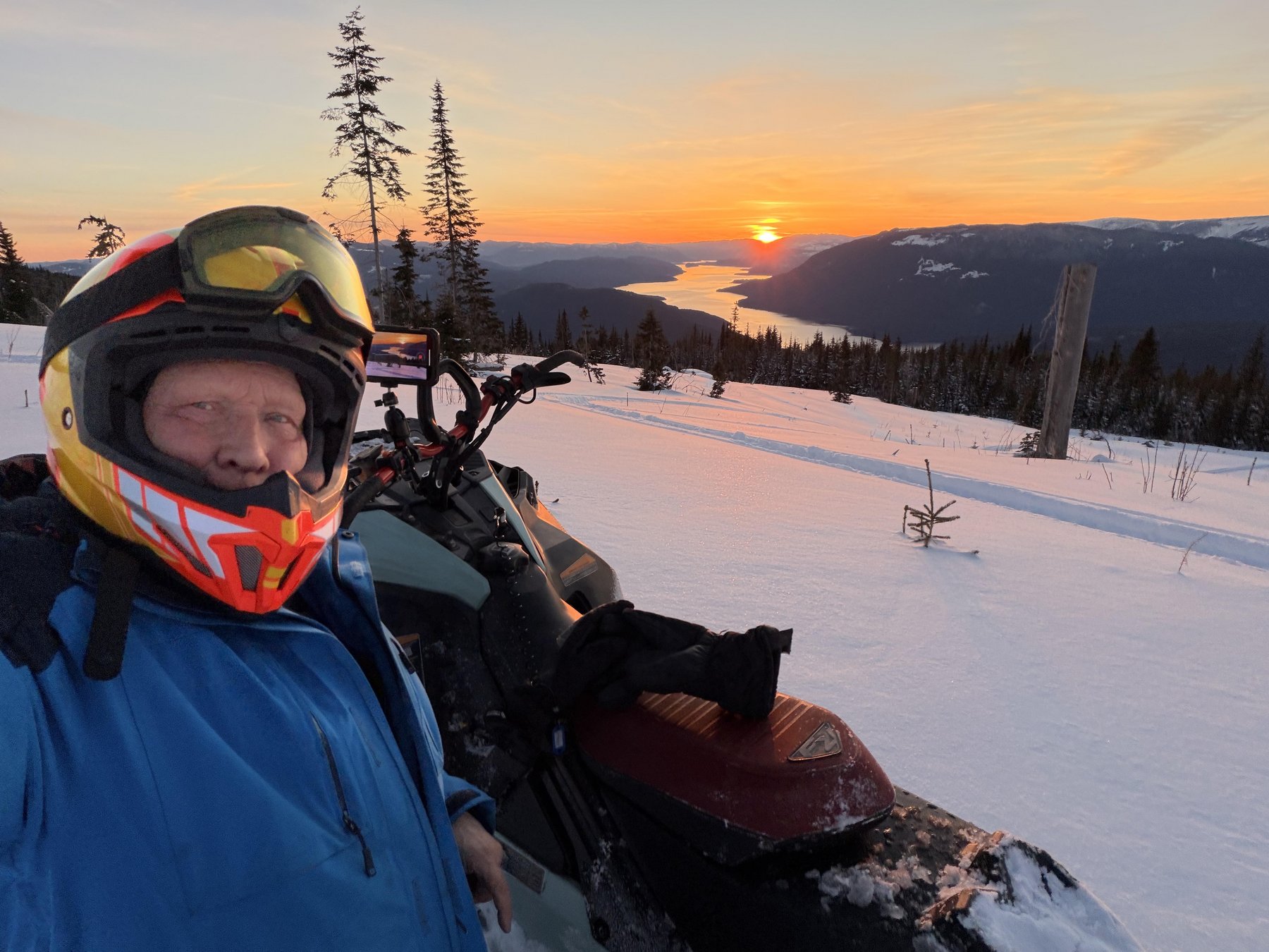 Snowmobile rider paused on a snowy ridge above Shuswap Lake as the sun sets behind the mountains