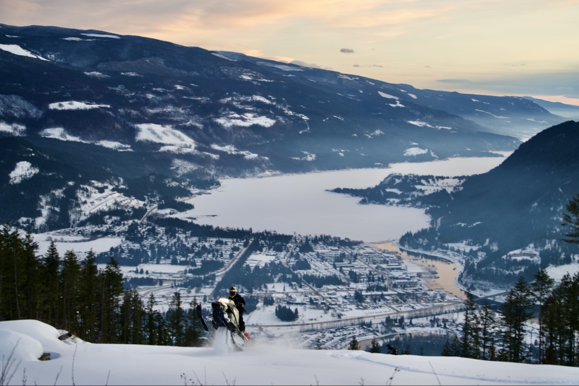Snowmobiler stopped on a snowy ridge overlooking Sicamous in winter, the frozen lake and snow-covered town spread out below