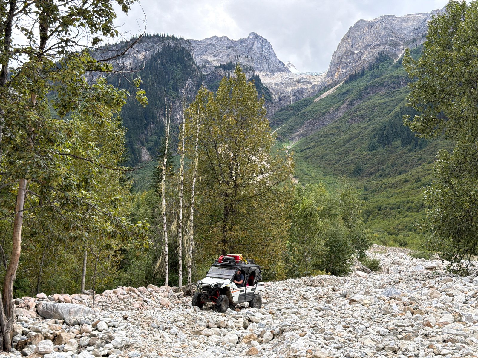 Side-by-side UTV on a rocky mountain trail among green trees with rugged peaks and a glacier behind