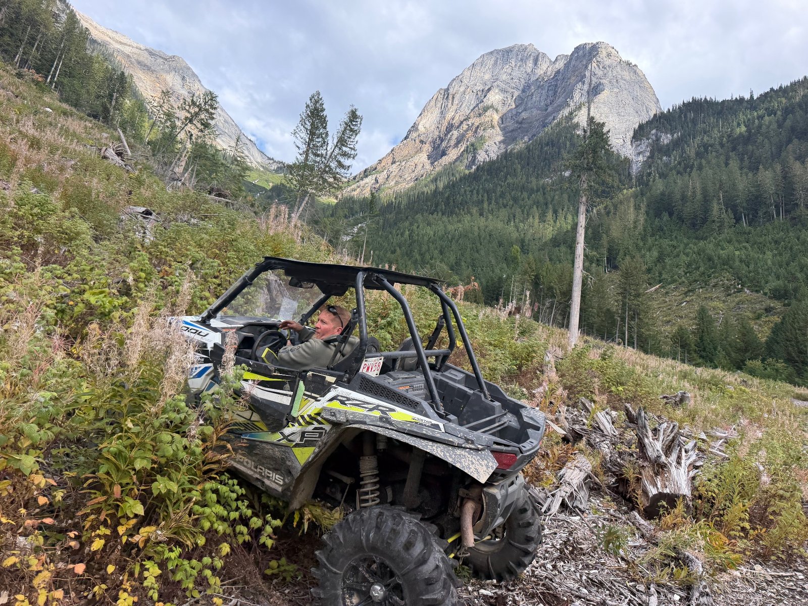 Polaris RZR side-by-side on a brushy mountain trail below forested slopes and a rocky peak