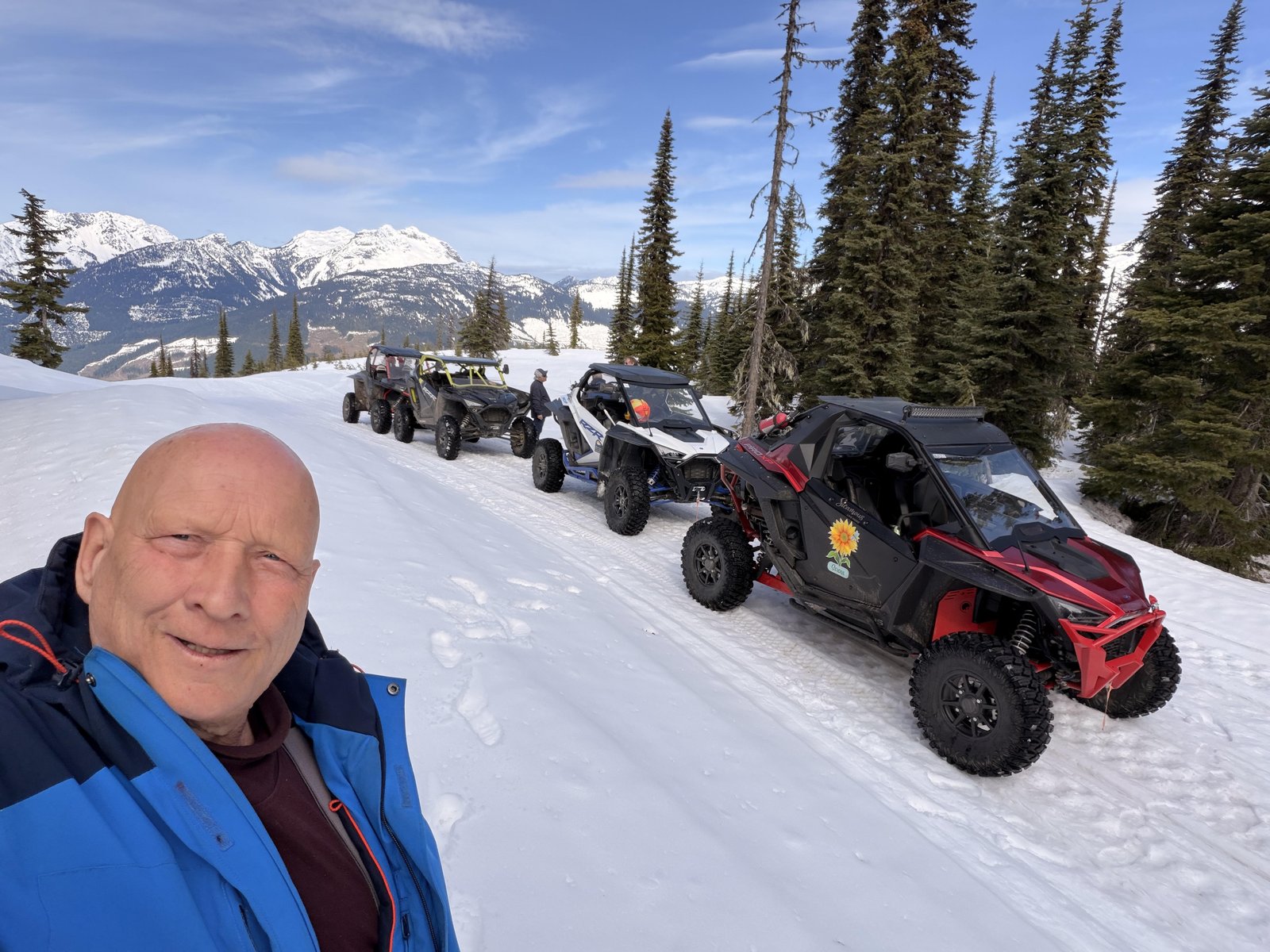 Red side-by-side parked on a snowy mountain trail with more side-by-sides lined up behind and snow-covered peaks in the distance