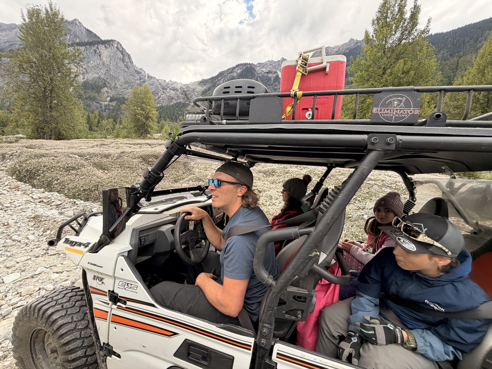 Side-by-side UTV carrying passengers on a rocky valley floor with green trees and rugged mountains behind in summer
