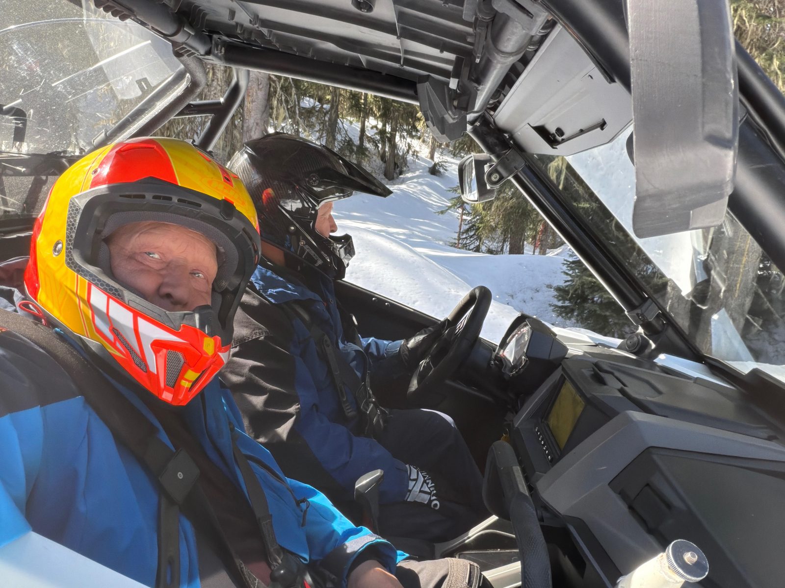 Two helmeted riders in a side-by-side UTV on a snowy forest trail, snow-covered trees outside the cab