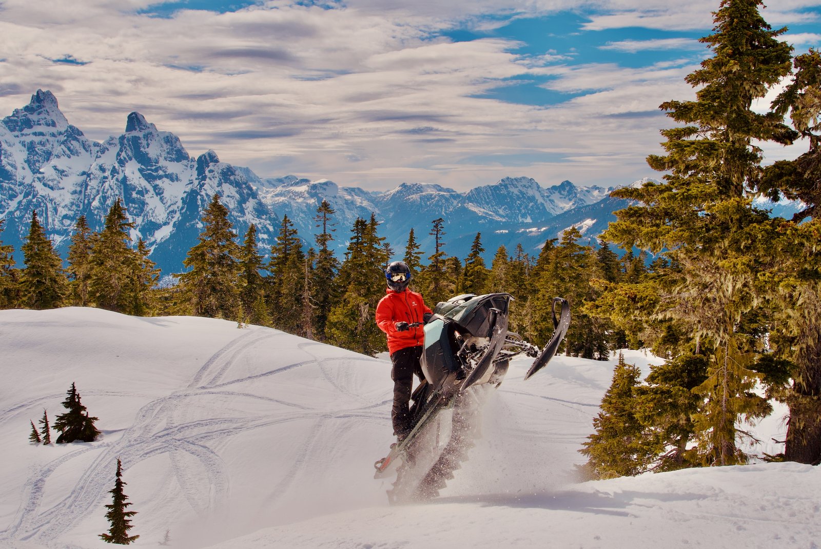 Snowmobile rider in a red jacket pulling a wheelie in deep powder below dramatic snow-covered peaks