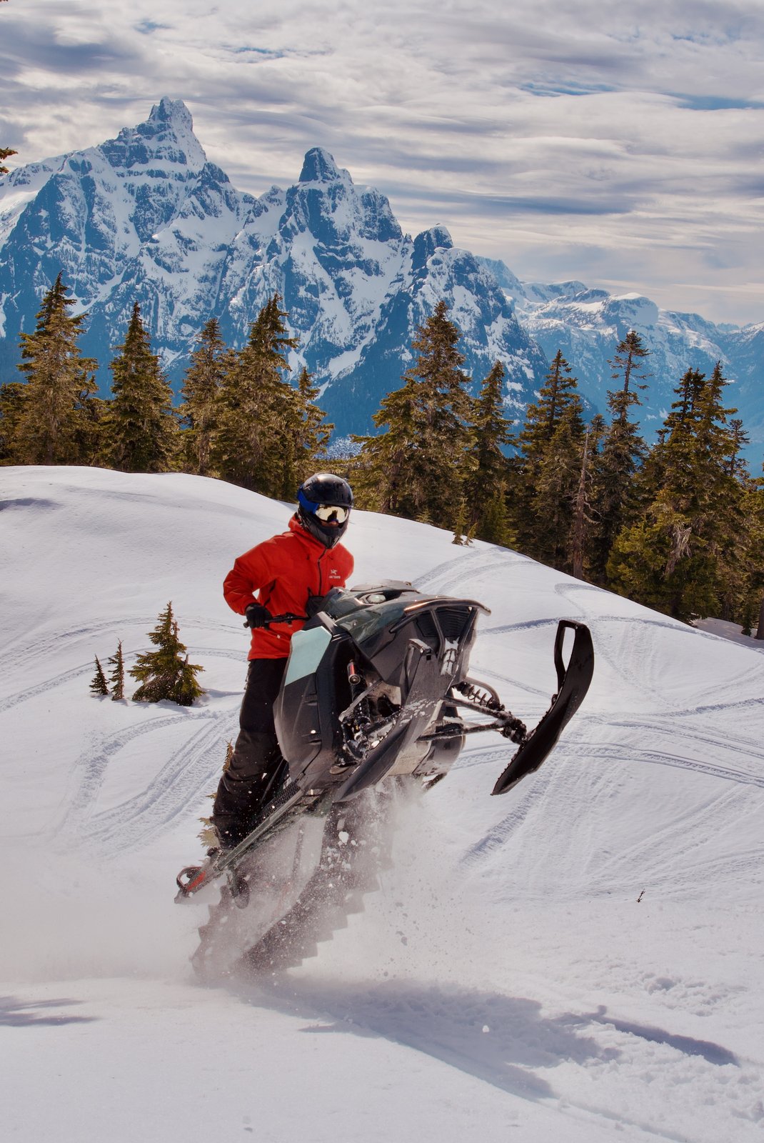 Snowmobile rider mid-jump with jagged snowy peaks behind