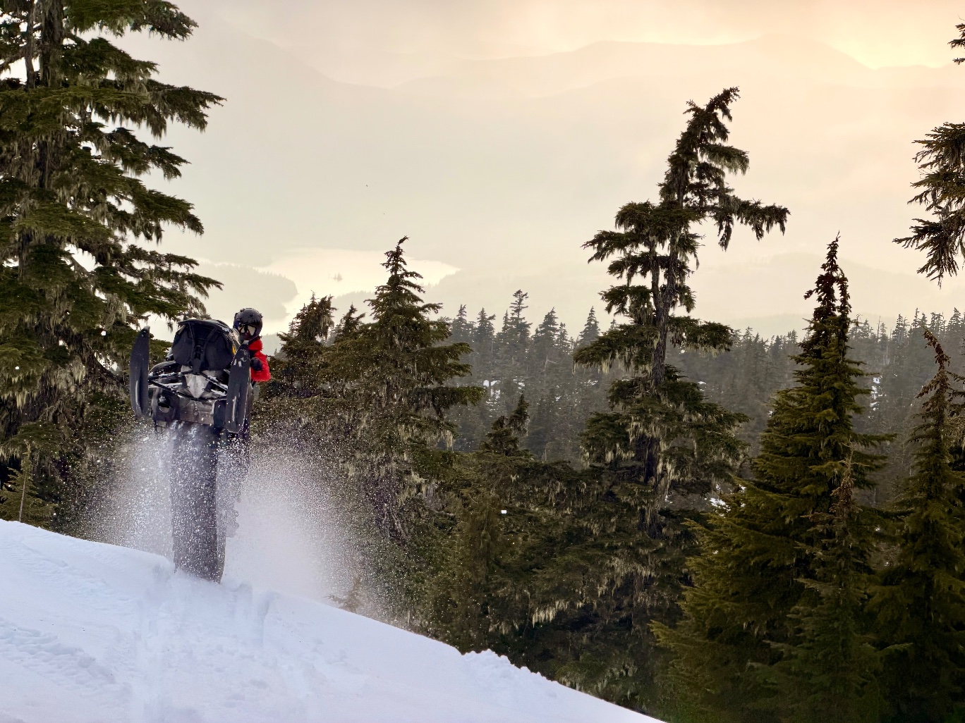 Snowmobile rider launching off a ridge at sunset above a forested valley