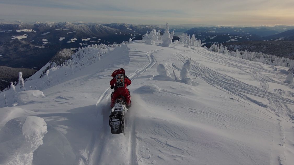 Snowmobile rider travelling along a snowy ridge with mountain ranges stretching to the horizon