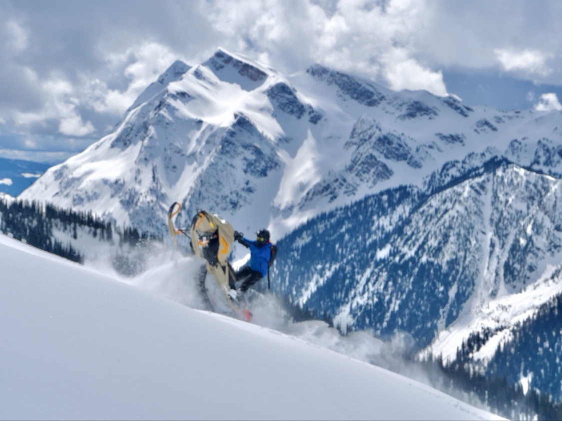 Snowmobiler catching air off a snowy ridge with big snow-covered peaks behind