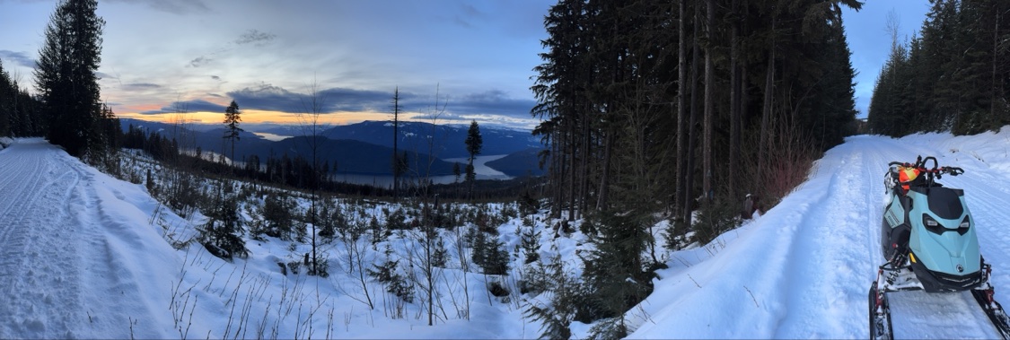 Panoramic winter sunset from a snowy snowmobile trail lined with evergreen trees, looking out over Shuswap Lake and the mountains